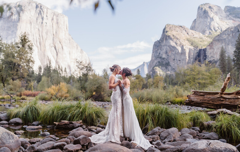 Two people in white wedding dresses embracing in a rocky riverbed with granite cliffs and evergreen trees in the background.