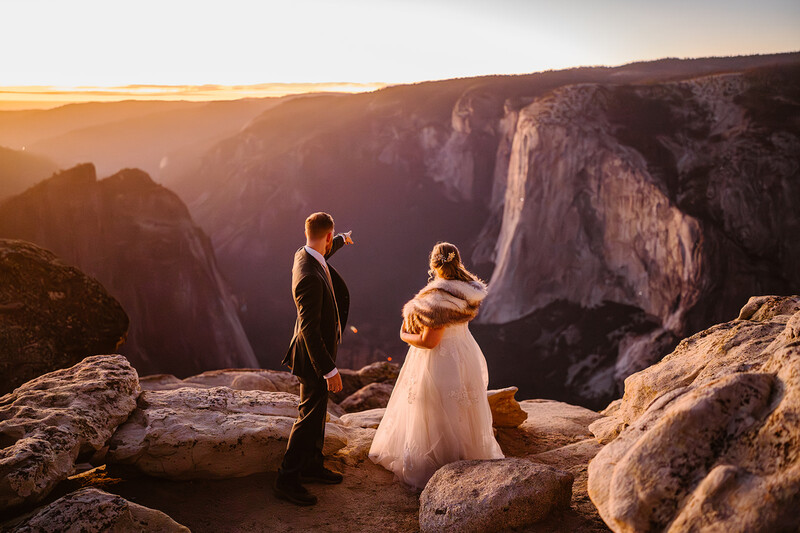 Bride and groom standing on rocky outcrop overlooking a vast canyon at golden hour.