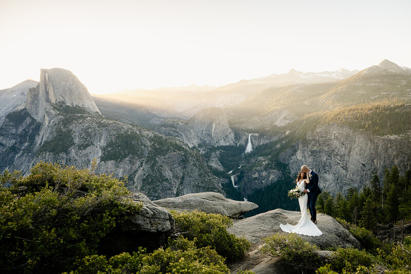 Bride and groom standing on a rocky overlook above a vast mountain valley at golden hour.