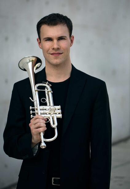 Young musician holding a trumpet against a neutral gray background.