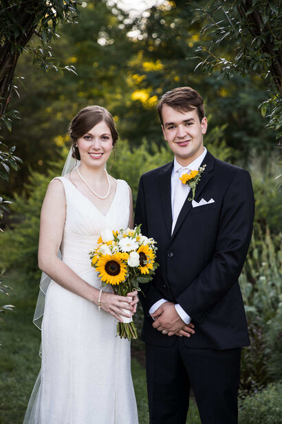 Bride and groom pose together outdoors holding a sunflower bouquet under tree branches.
