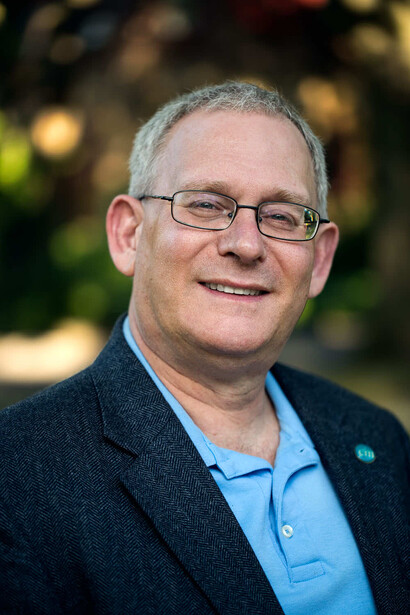 Portrait of a smiling man wearing glasses and a light blue shirt under a dark blazer.