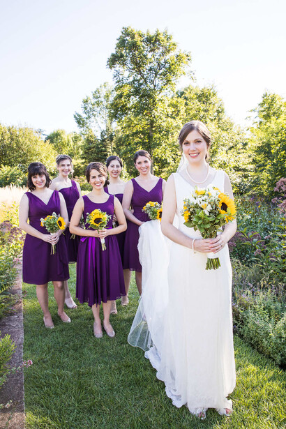Bride in white dress with bridesmaids in purple dresses holding yellow bouquets in garden setting.