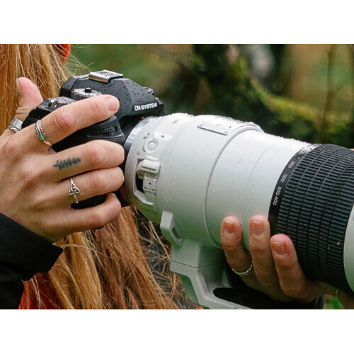 Photographer holding a DSLR camera with telephoto lens, wearing multiple rings and bracelets.