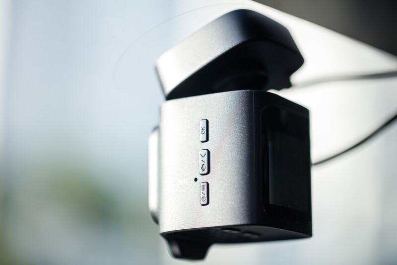 Compact dashcam mounted on vehicle windshield with silver and black finish and three control buttons visible.