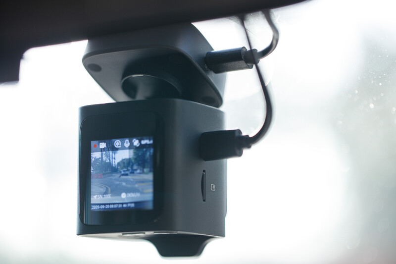 Dashboard camera mounted on vehicle windshield displaying live street view on small screen.