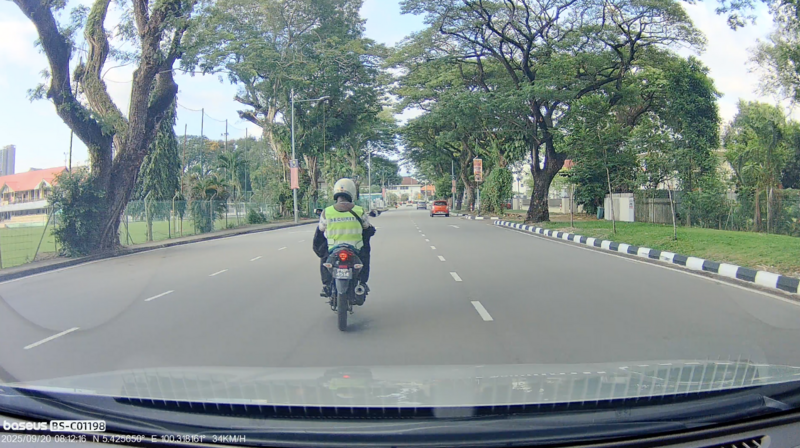 Motorcyclist in yellow safety vest riding ahead on tree-lined asphalt road, shot from inside a vehicle.