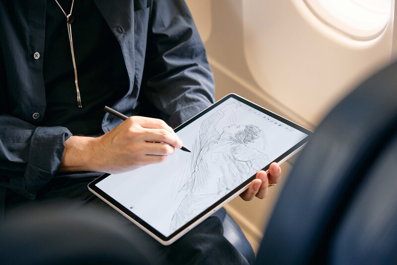 Person in dark jacket holding tablet device with sketch drawing while seated by airplane window.