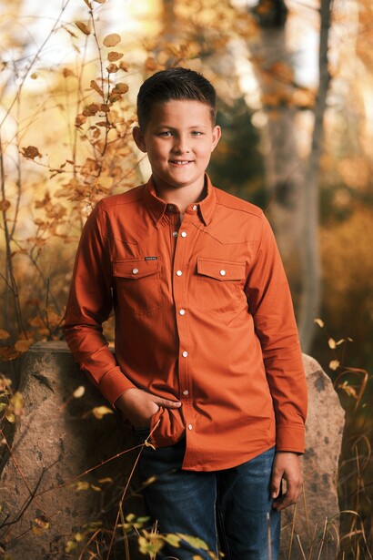 Young boy in orange shirt standing among dried autumn plants and branches.