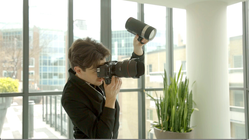 Photographer holding DSLR camera with telephoto lens to eye, positioned at floor-to-ceiling windows overlooking city skyline.