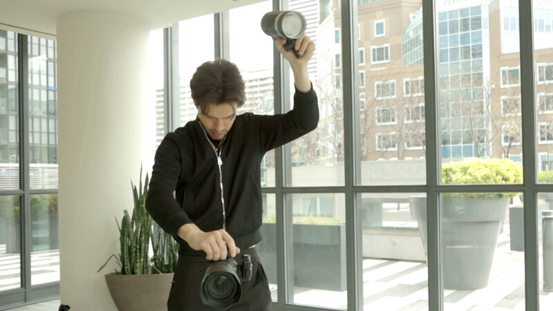 Photographer holding light meter and camera in bright window studio with city skyline behind.