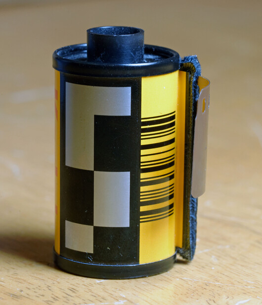Film canister with yellow and black checkered pattern and blue top against neutral background.