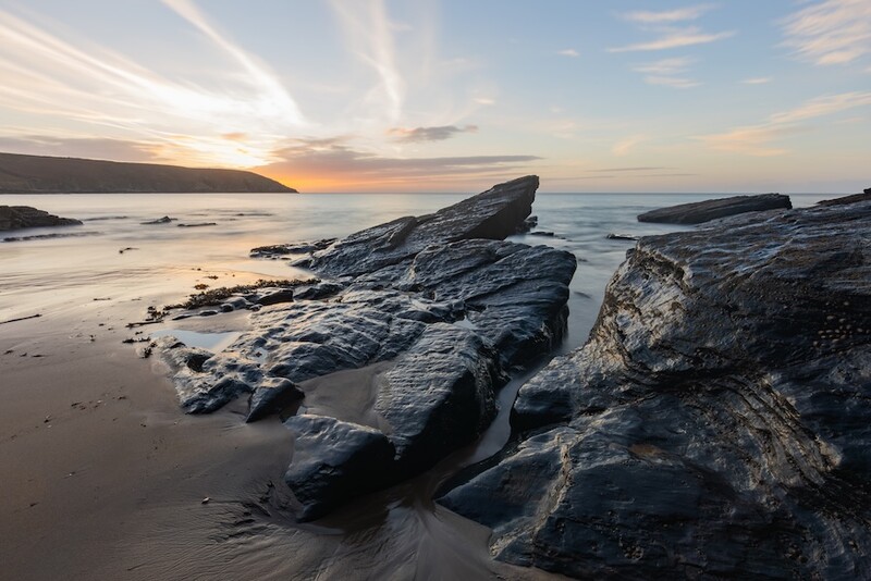 Jagged dark rock formations jutting from wet sand at sunset, with golden sky and calm sea horizon.