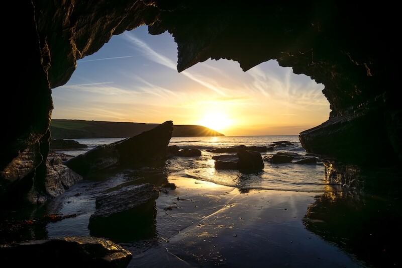 Sunset viewed through a cave opening, with golden light reflecting off wet sand and rocks.