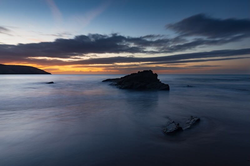 Rocky island silhouetted against golden sunset sky with smooth water and distant coastline.