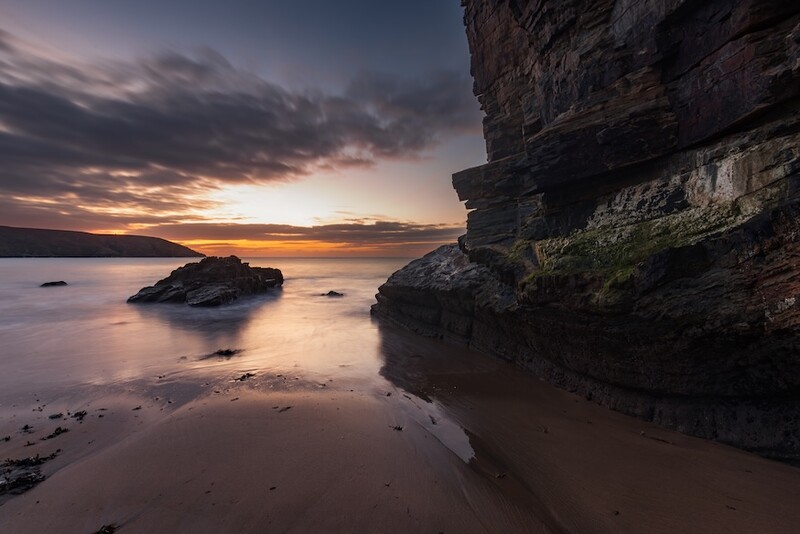 Rocky coastline at sunset with calm waters and a small island in the distance.