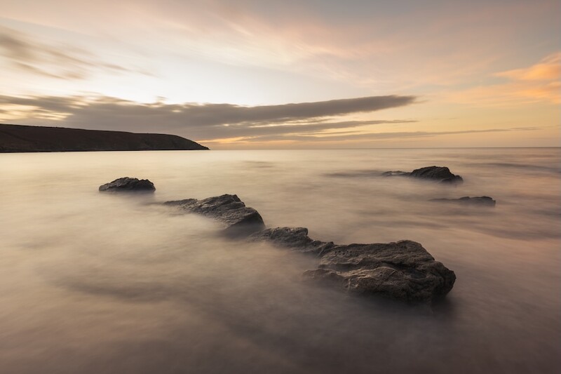 Rocky outcrops emerging from smooth, misty seawater at twilight with a headland silhouetted against a golden-pink sky.