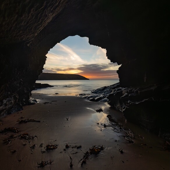 Sunset viewed through a sea cave opening, with rocky beach and wet sand in foreground.