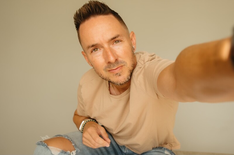 Man in tan t-shirt taking a selfie against neutral background.