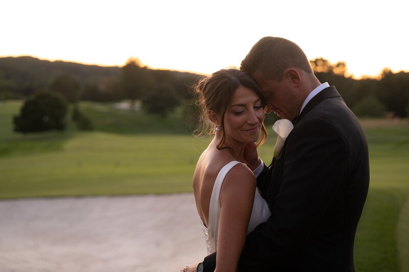 Bride and groom embracing outdoors at golden hour with green fields in background.