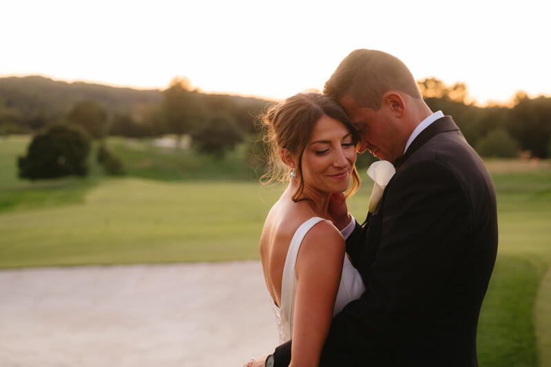 Bride and groom posing together outdoors during golden hour with green fields behind them.