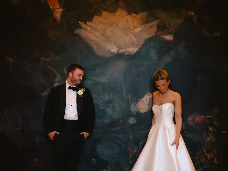 Bride and groom posing together against a large painted landscape backdrop in formal wedding attire.