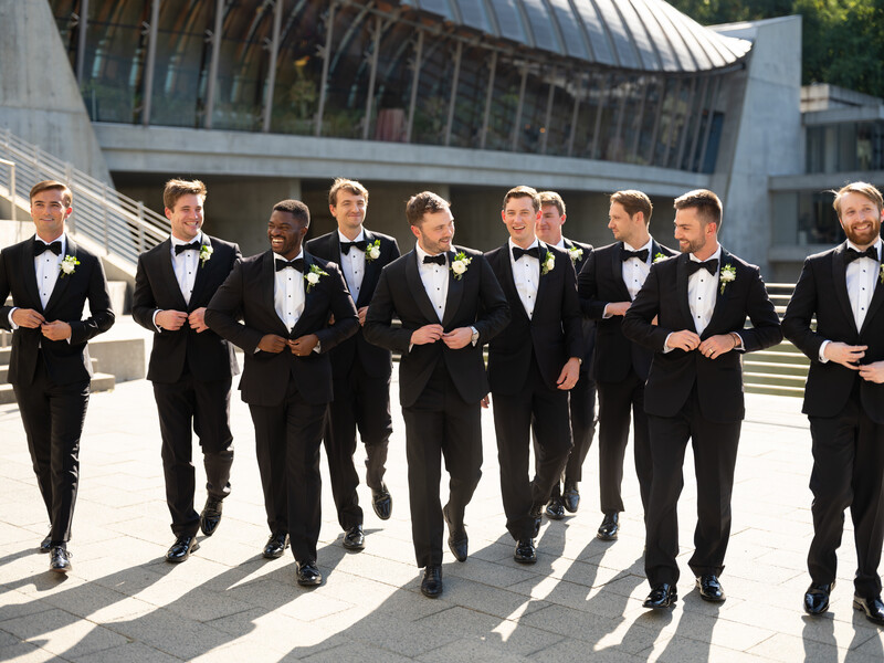 Nine men in black tuxedos walking together across a paved plaza in bright sunlight.