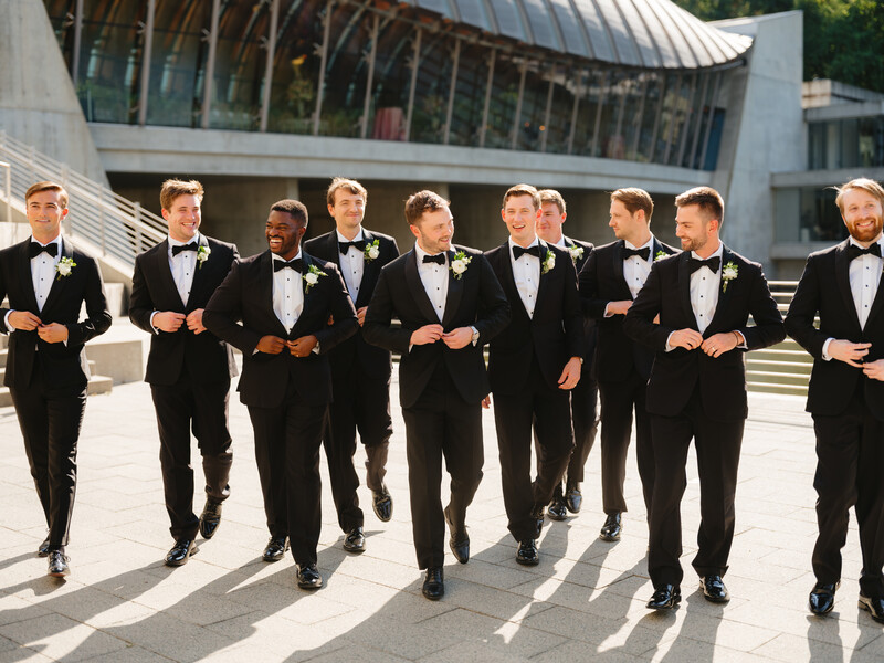 Nine men in black tuxedos walking in formation across a sunny plaza with modern architecture behind them.