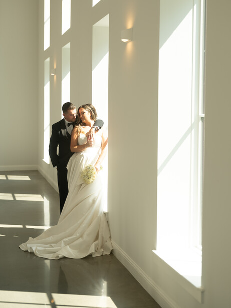 Bride and groom embrace in a bright hallway with floor-to-ceiling windows casting dramatic shadows.