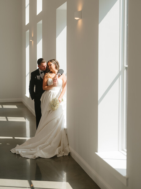 Bride and groom embracing in a bright hallway with floor-to-ceiling windows casting dramatic sunlight.
