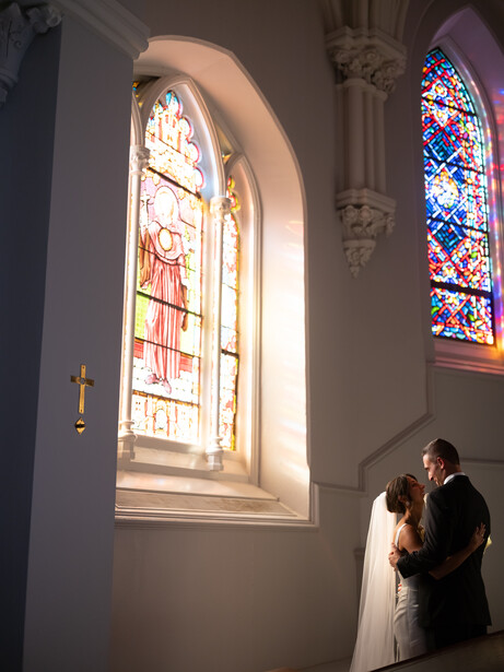 Bride and groom standing beneath illuminated stained glass windows in a church.