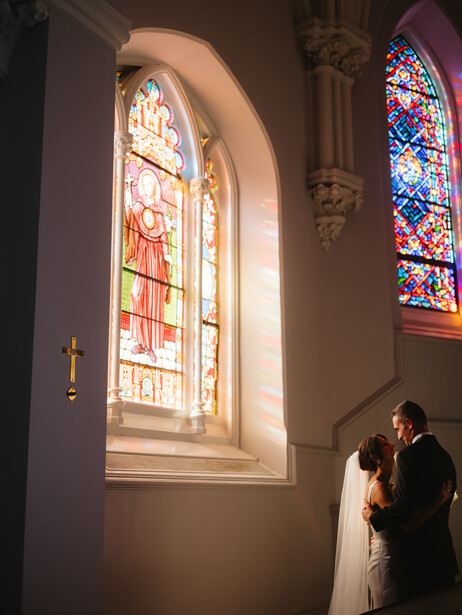 Bride and groom standing before an illuminated stained glass window in a church.