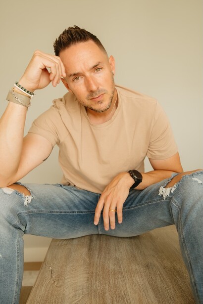 Man in tan t-shirt and jeans sitting on wooden floor against neutral background.