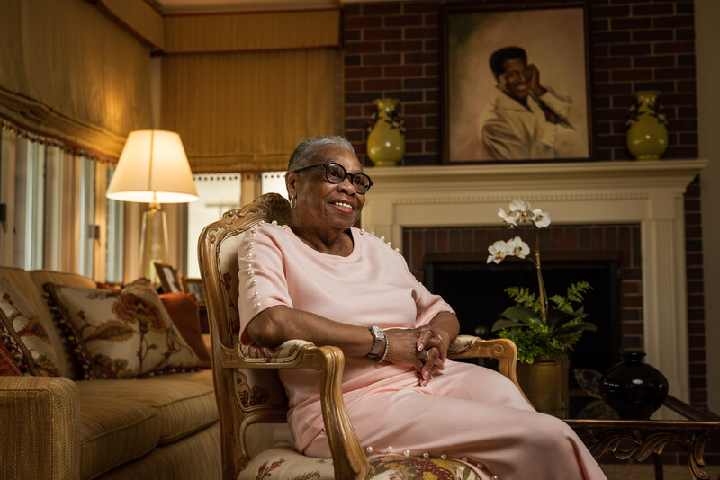 Older woman with glasses seated in an upholstered chair in an elegant living room with fireplace and artwork.