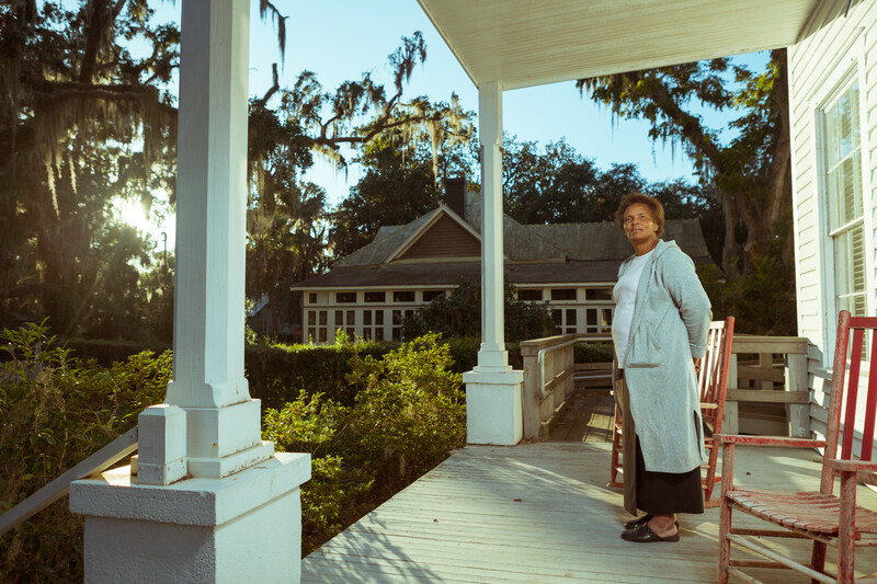 Woman standing on a covered porch with white columns, surrounded by trees and Spanish moss.