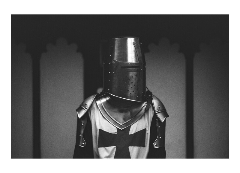 Medieval knight in full armor and helmet stands facing the camera in a stone corridor.