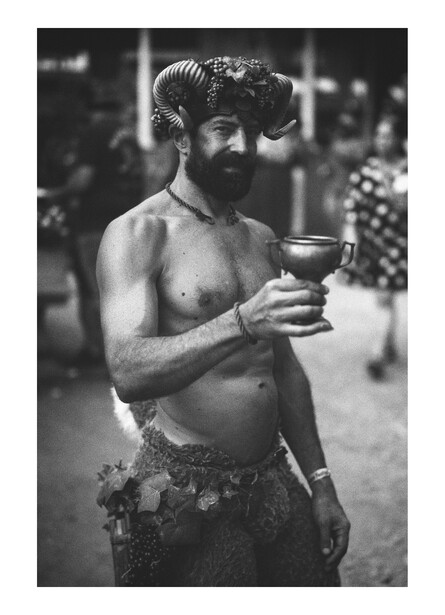 Shirtless man with beard holding cocktail glass, shot in black and white with blurred background.