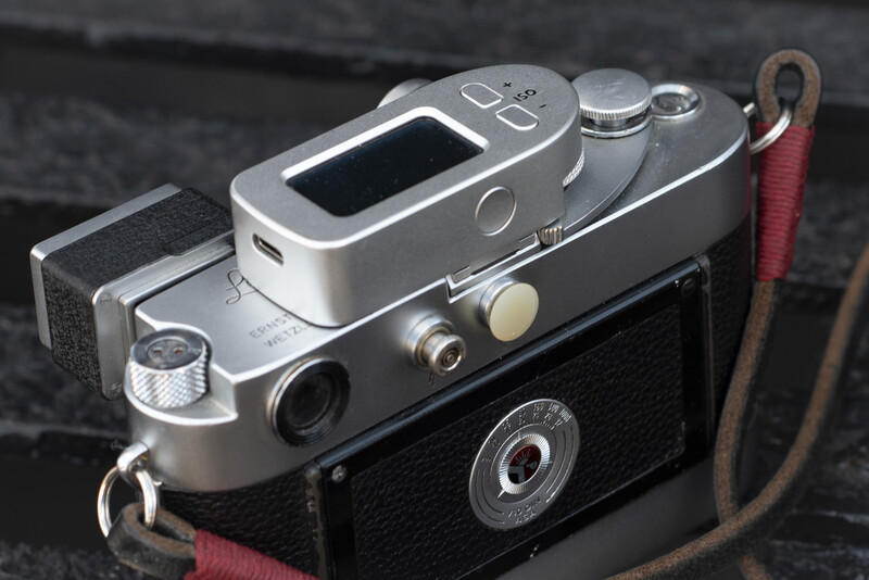 Vintage rangefinder camera with silver top plate and black body, displayed with a red fabric strap against a dark surface.