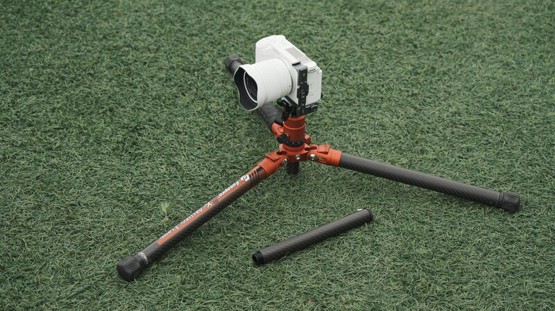 White compact camera mounted on red and black tripod legs against green textile backdrop.