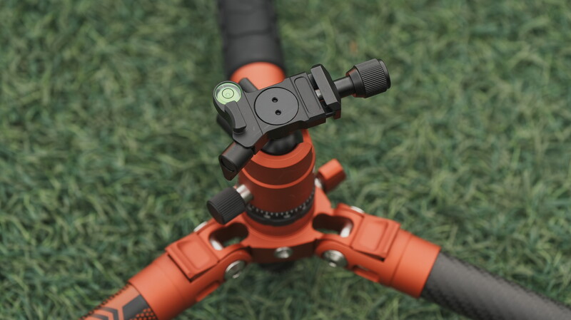 Red tripod head with ball joint and adjustment knobs mounted on black legs against artificial green grass background.