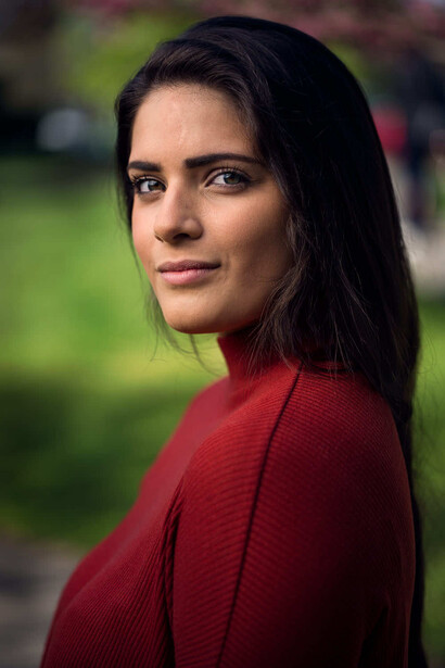 Woman with long dark hair wearing a red sweater, photographed outdoors with a blurred green background.