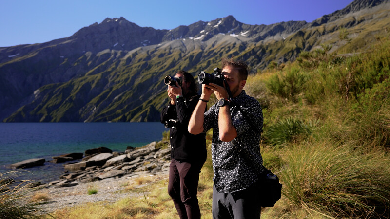 Two photographers with binoculars and telephoto lenses observing wildlife beside an alpine lake with mountains beyond.