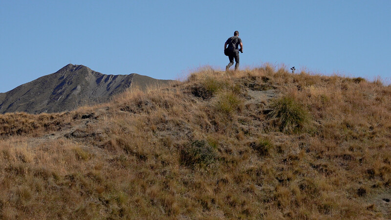 Hiker ascending a sparse, arid hillside dotted with dry grass and volcanic rock under clear blue sky.