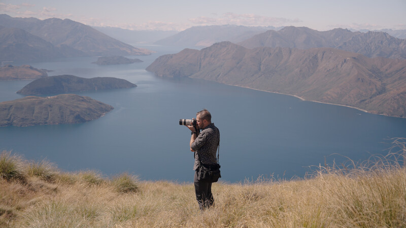 Photographer with telephoto lens standing on hillside overlooking expansive alpine lake and mountains.