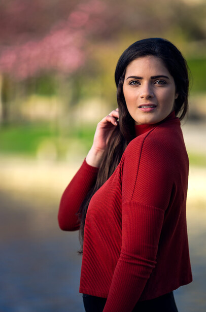 Woman in red cardigan looking over shoulder toward camera with soft outdoor background.