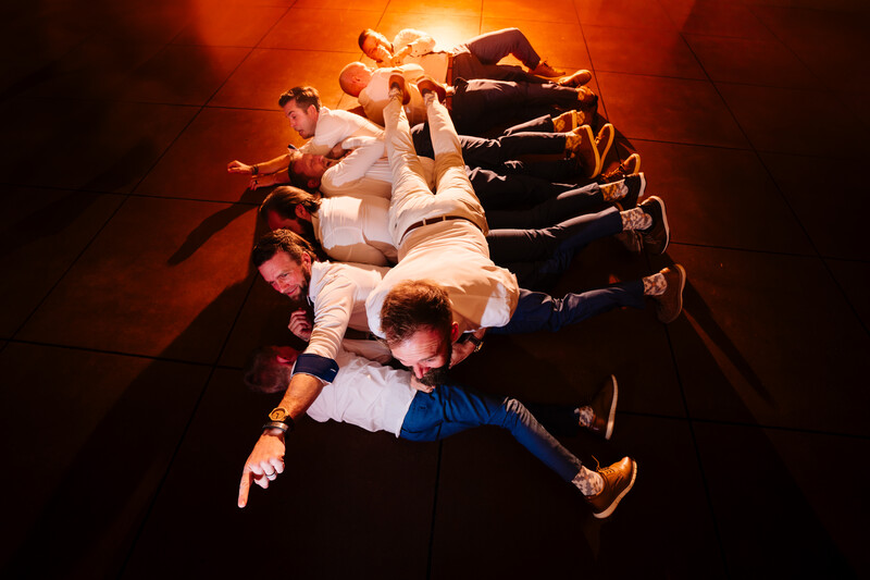Group of people lying on dark floor arranged in circle, viewed from above with warm orange lighting.