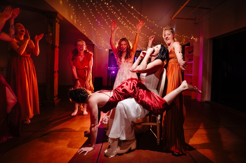 Woman in white dress being lifted by man in suit during energetic party scene with dancing guests in warm red and orange lighting.