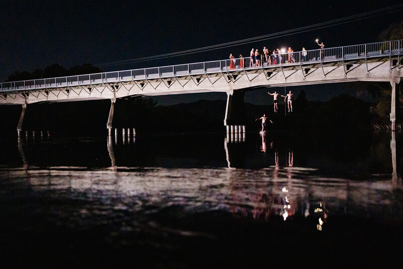 People standing on an illuminated bridge at night reflected in still water below.