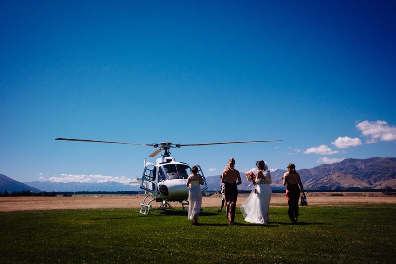 Wedding party standing beside a helicopter on a grass field with mountains in the background.