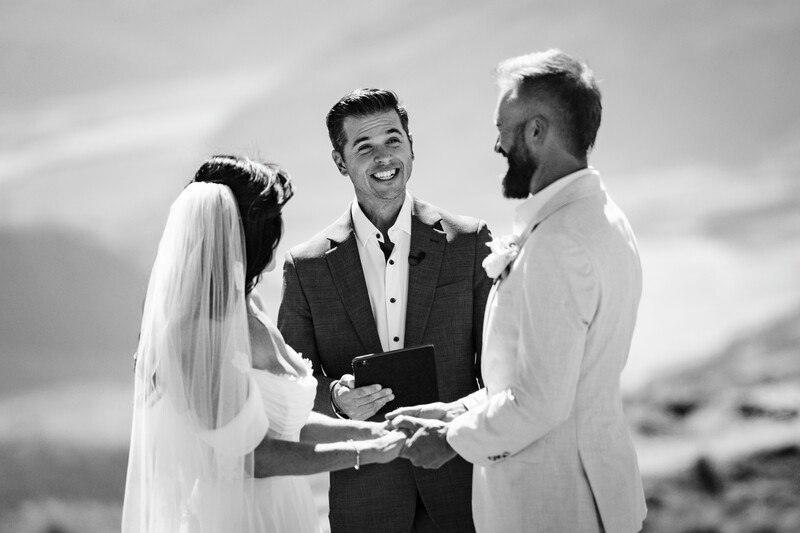 Officiant in dark suit smiling while conducting wedding ceremony between bride in white dress and groom in white shirt.